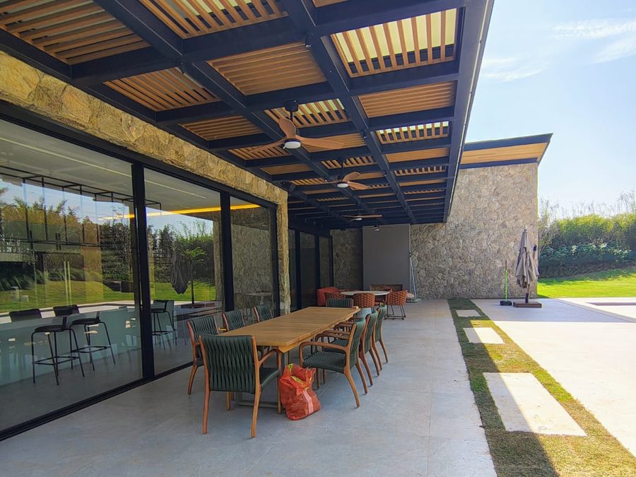 Varanda gourmet de residência de alto padrão com pergolado metálico e ripado de madeira, fechamento em vidro e parede de pedra natural